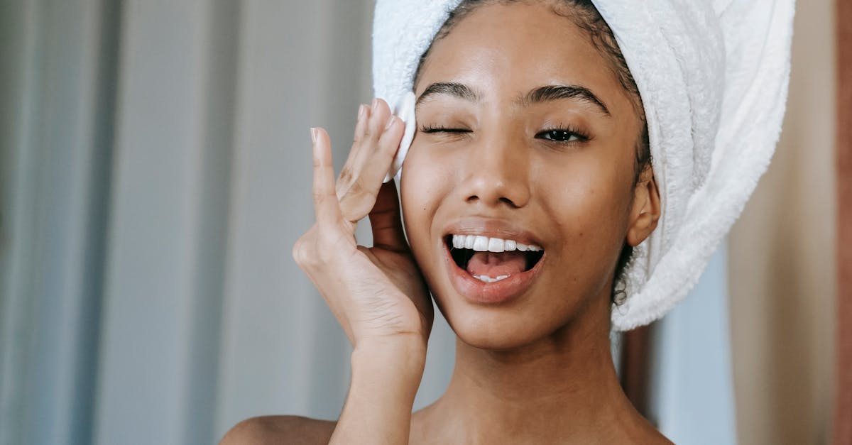 Young woman with towel on head, smiling and applying skincare product, promoting morning beauty routine.