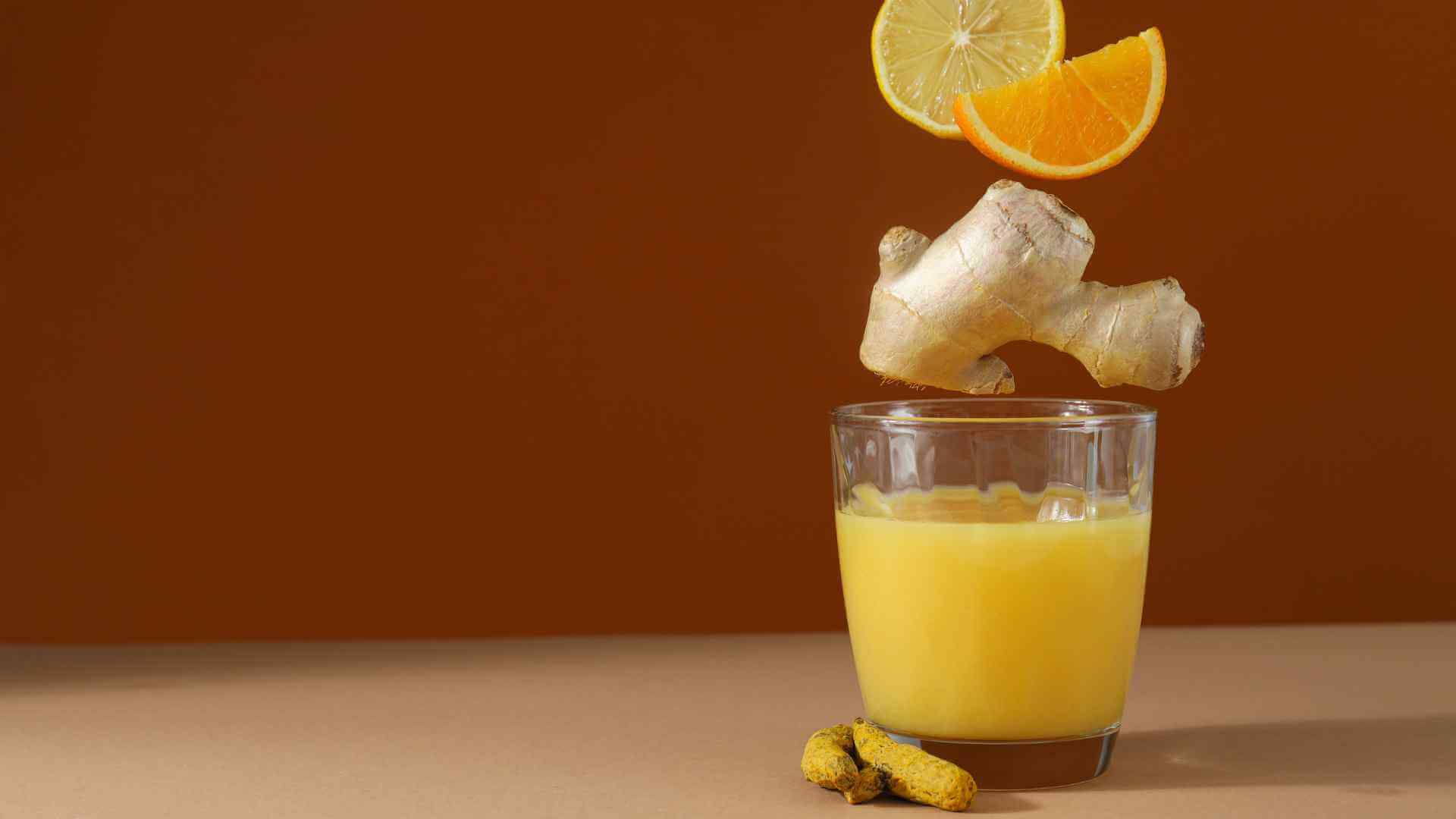 A dynamic composition featuring a glass of turmeric ginger lemonade, with floating ginger, lemon, and orange slices above, set against a rich brown backdrop.