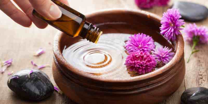 and pouring essential oil into a wooden bowl with floating flowers for aromatherapy infusion