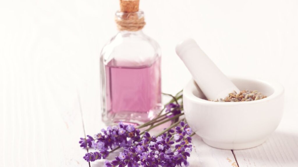 Bottle of lavender essential oil beside fresh lavender flowers and a white mortar on a wooden table.