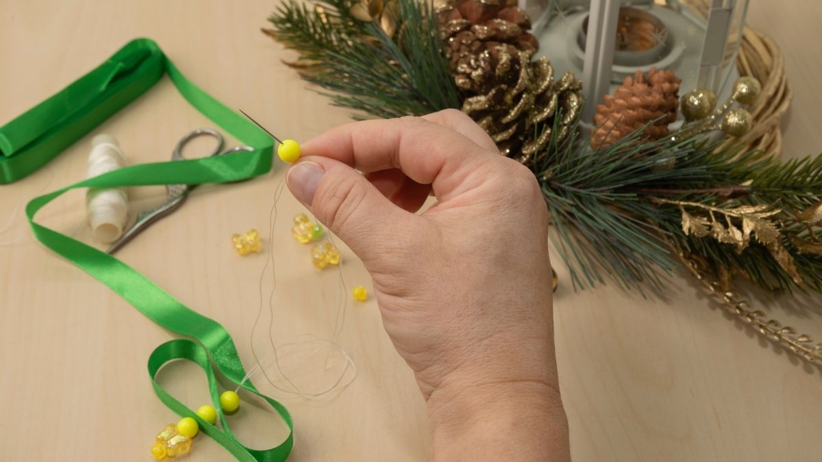 A hand threading yellow beads onto a needle beside green ribbon, small craft supplies and evergreen Christmas decorations on a table.