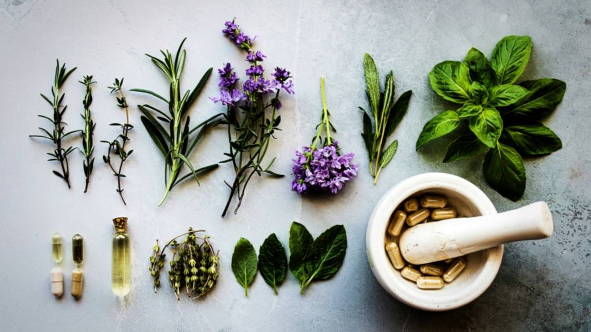 Fresh herbs, flowers, capsules and a mortar and pestle arranged on a light surface, showing ingredients commonly used in natural wellness.