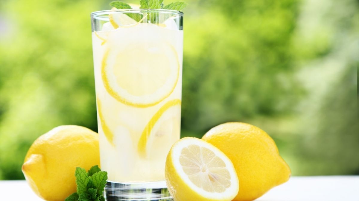 A glass of lemon water with fresh lemon slices and whole lemons on a table outdoors.